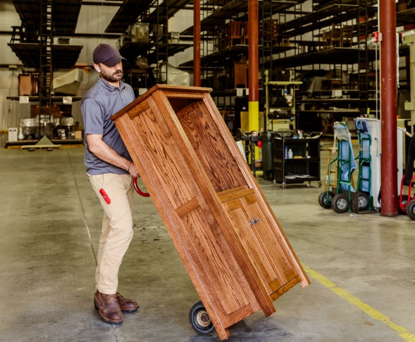 A warehouse worker in a purple cap and gray polo shirt uses a hand truck to transport a large wooden door through an industrial warehouse facility with metal shelving and equipment visible in the background. This is a Millwest employee preparing a piece of furniture for a delivery.