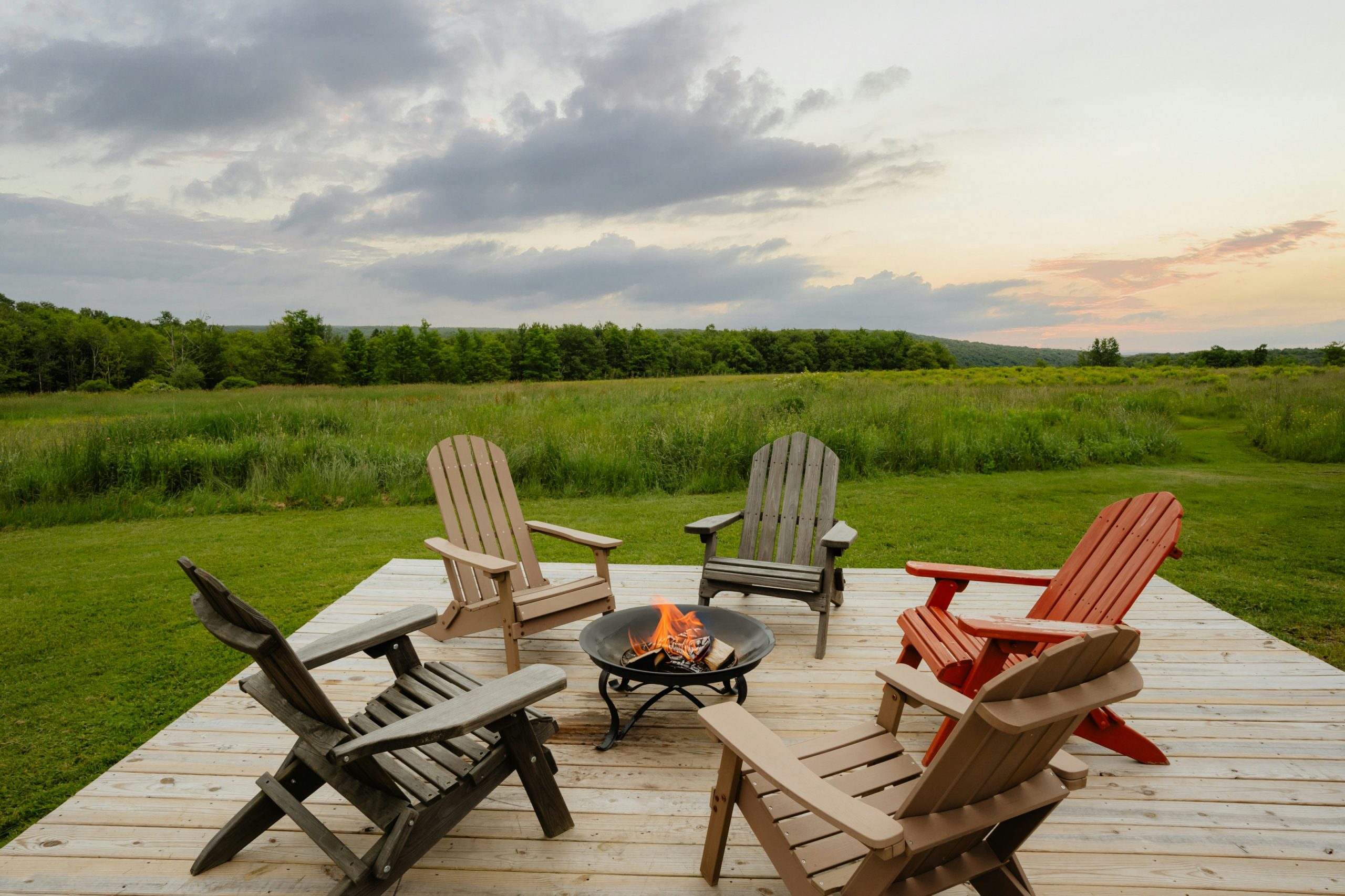 Adirondack-style wood and poly outdoor chairs around a fire pit on a wooden deck, set against a scenic countryside — Millwest Amish Furniture blog on choosing between wood and poly furniture