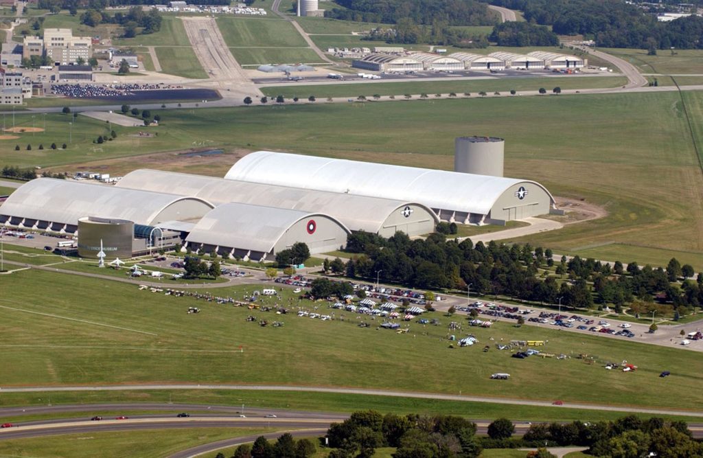 Aerial view of a large aviation facility featuring multiple curved-roof aircraft hangars with US Air Force insignia, situated alongside an airport runway. The complex includes several massive white hangar buildings, a modern visitor center or museum building, and extensive parking areas filled with cars. The facility is surrounded by green agricultural fields and wooded areas, with additional airport infrastructure visible in the background including runways and terminal buildings. This museum is located near Fairborn, where Millwest delivers Amish furniture.