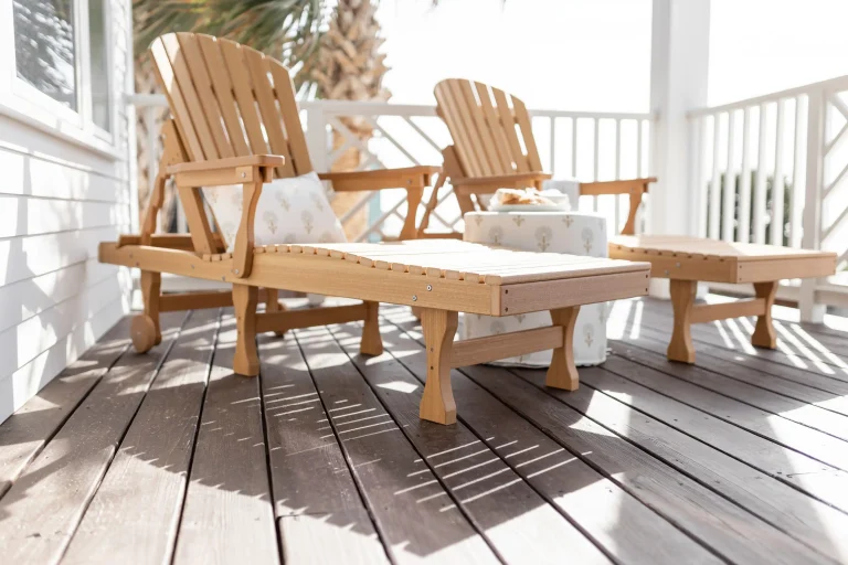 Two tan poly lumber Adirondack chairs with white cushions and matching ottoman on a sunny beach house deck