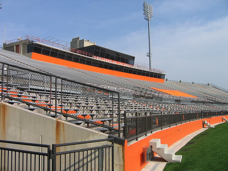 Empty football stadium with aluminum bleacher seating and orange accents. The stadium features a concrete structure with metal railings, a press box with 'WELCOME TO THE DOYT' signage, stadium lighting, and artificial turf field visible in the foreground. This is Doyt Perry Stadium at Bowling Green State University in Bowling Green, where Millwest provides furniture.