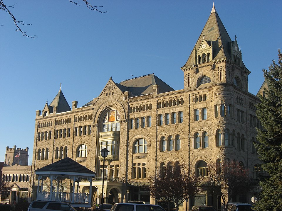 A grand Victorian-era stone building showcasing Romanesque Revival architecture, featuring a prominent corner tower with a conical roof, elegant arched windows, intricate stonework, and multiple stories. This historic landmark, Fort Piqua Plaza, stands as an architectural centerpiece in a community served by Millwest furniture suppliers.