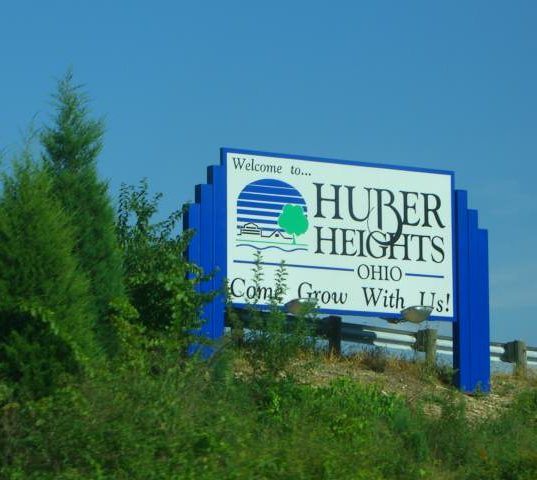 A blue and white welcome sign for Huber Heights, Ohio, where Millwest provides quality Amish furniture. The sign reads 'Welcome to... HUBER HEIGHTS OHIO Come Grow With Us!' and features a logo with green trees and blue water elements. The sign is positioned alongside a road with lush evergreen trees and vegetation in the background under a clear blue sky.