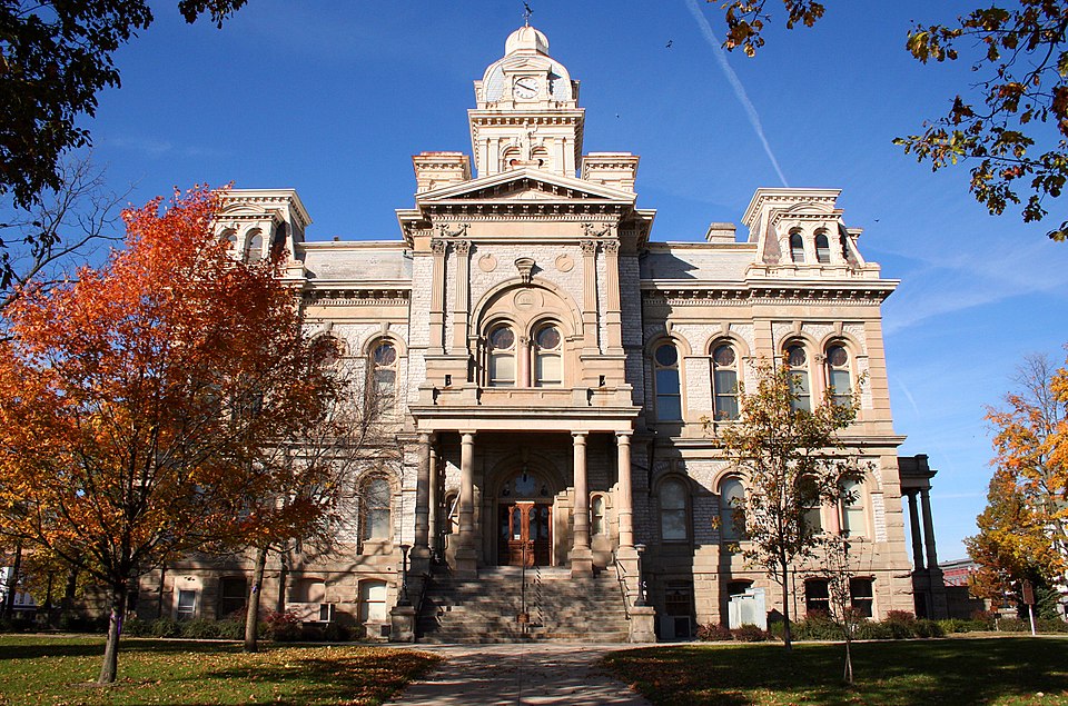 A grand Victorian-era courthouse with ornate limestone architecture featuring a central clock tower, arched entrance with columns, and symmetrical wings. The building is surrounded by autumn trees with vibrant orange and red foliage against a clear blue sky in the beautiful town of Sidney, where Millwest offers furniture.