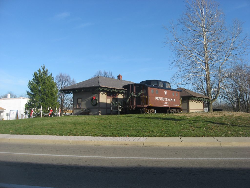 A vintage Pennsylvania Railroad caboose numbered 477130 sits on display next to a historic train depot building. The maroon and tan caboose features the classic cupola design and Pennsylvania Railroad lettering. The depot is a single-story structure with decorative trim and a brick chimney. Both are positioned on a grassy area with mature bare trees in the background under a clear blue sky. Holiday wreaths and decorations are visible on the depot building. The scene is viewed from across a paved road at this preserved railway heritage site or museum display in Trotwood, where Millwest delivers furniture.