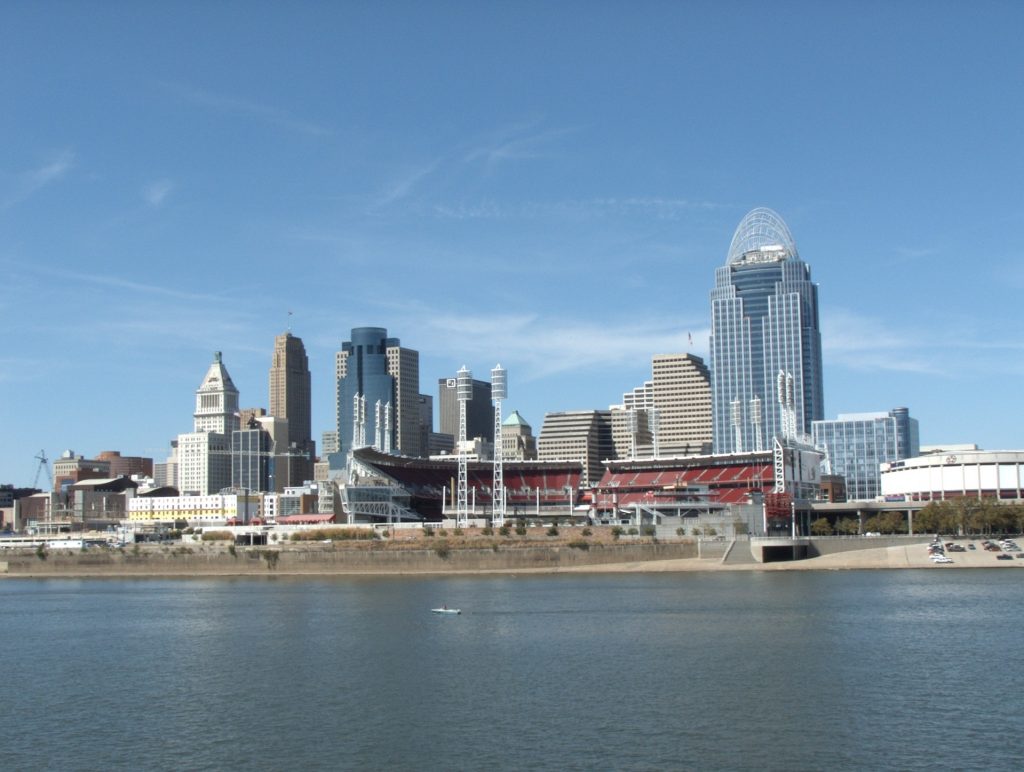 Cincinnati skyline viewed from across the Ohio River on a clear day, featuring a baseball stadium with red seating in the foreground and downtown skyscrapers rising behind it, including a prominent tall building with an illuminated crown-like architectural feature at the top. Millwest provides quality Amish furniture to this city.