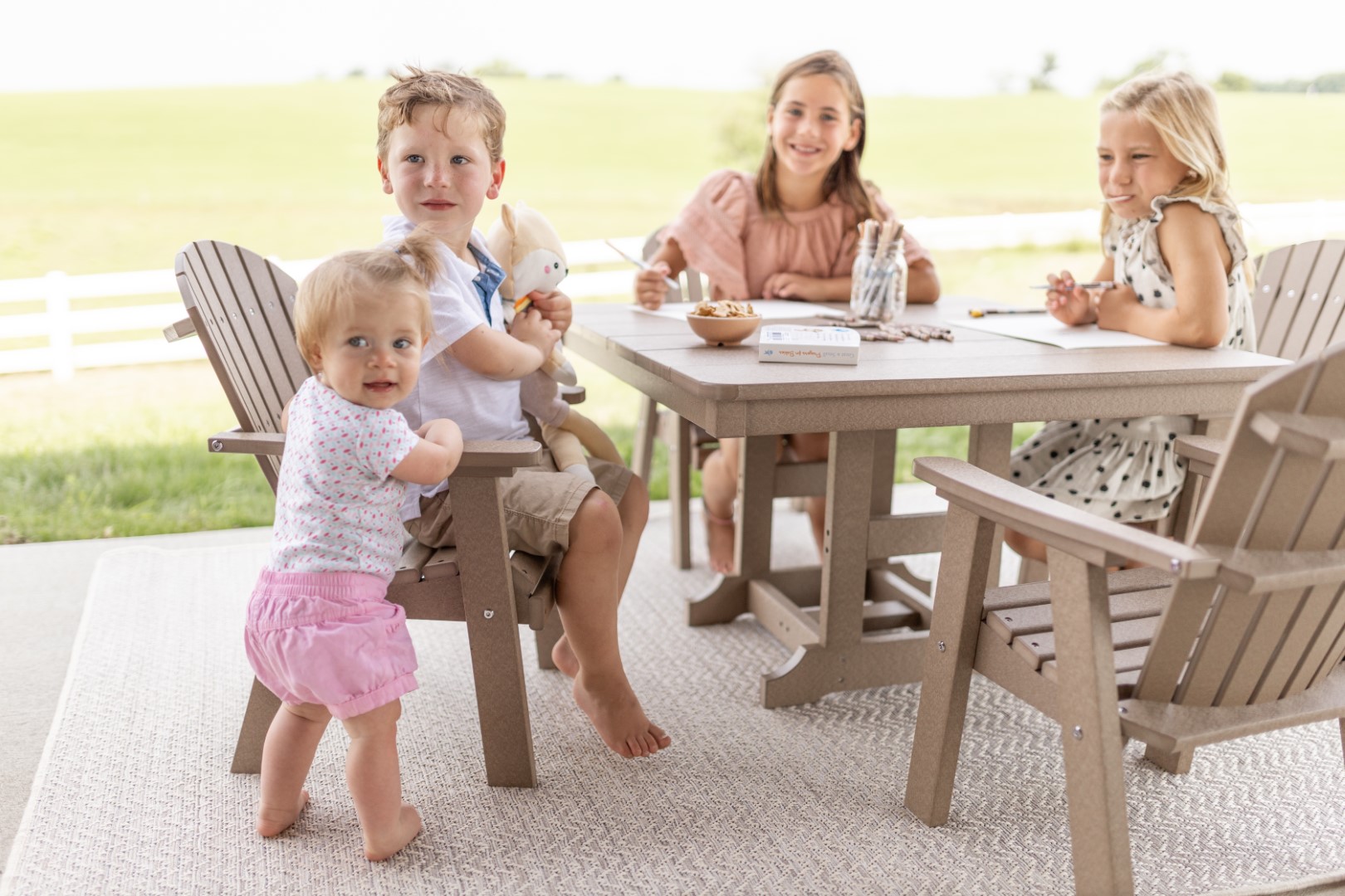Four children enjoying snacks at a poly lumber outdoor table on a covered patio. A toddler in a pink skirt stands in front while an older boy sits in an Adirondack chair behind her. Two girls sit at the table with bowls and craft supplies, with green pastoral fields visible in the background.