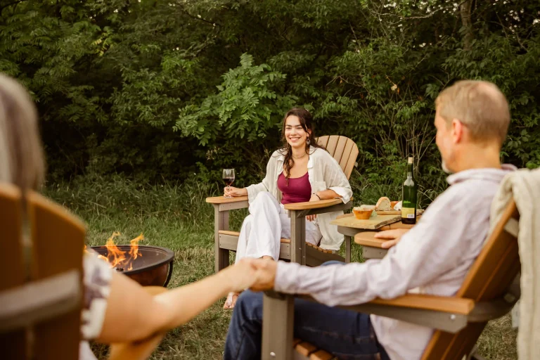 Friends relaxing in poly lumber Adirondack chairs around a fire pit, enjoying wine and snacks in a lush backyard setting